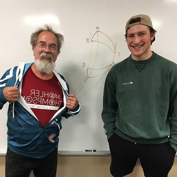 一个微笑的学生和教授站在一块写着图表的白板前. 教授拉开连帽衫的拉链，露出了他的Mohler Thompson AQ夏季研究T恤. 