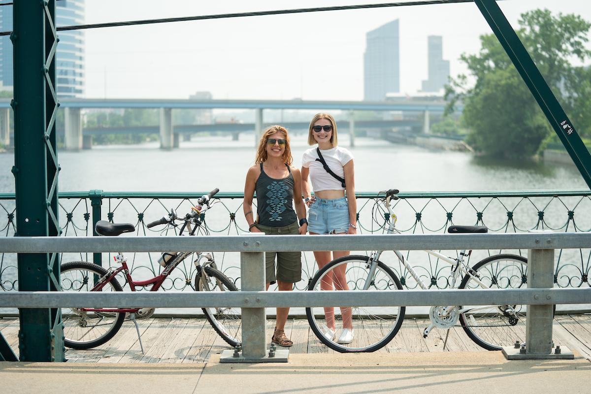 Dr. 伦德鲁姆和阿拉贝拉·卡明斯骑着自行车站在大急流城的一座桥上, 背景是河流和城市景观